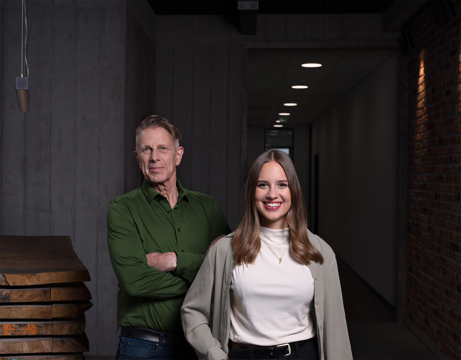 Milena und Jochen Greim, Inhaber des familiengeführten Unternehmens Die Greim Gruppe stehen in einem modernen Korridor mit Backsteinwand und Holzplatten, symbolisierend die Verlässlichkeit und langfristigen Partnerschaften, die das Unternehmen in vierter Generation auszeichnet.