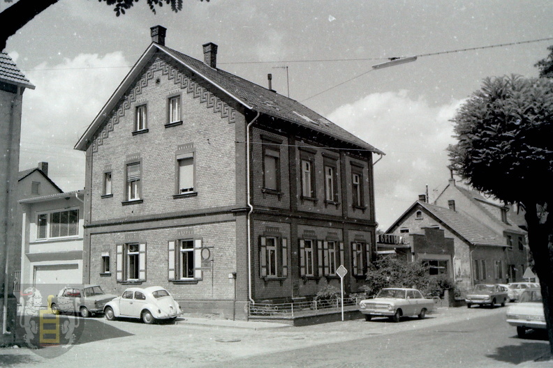 Schwarz-Weiß-Foto eines zweistöckigen Ziegelhauses mit Parkplatz und klassischen Autos an einer Straße in Wörrstadt, Rheinland-Pfalz, früherer Standort der Greim Gruppe.