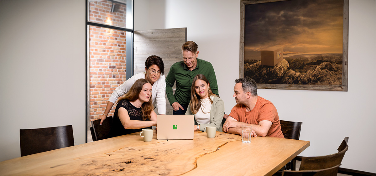 Ein Team der Greim Gruppe kooperiert in einem modernen Büro an einem Holztisch an einem Laptop—das Bild veranschaulicht die familiäre Unternehmenskultur und die praxisorientierten Dienstleistungen in Fulfillment, Logistik und Consulting.