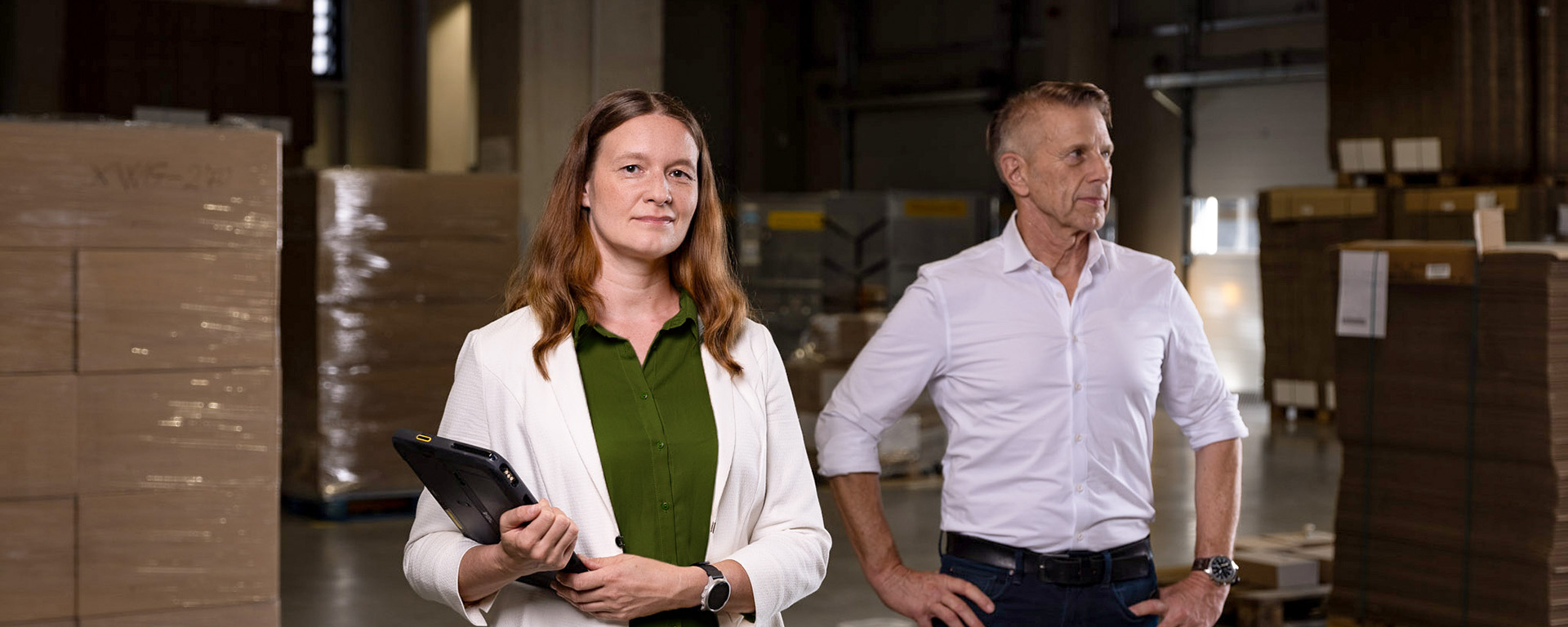 Christina Richter im Vordergrund und Jochen Greim im Hintergrund, der Greim Gruppe in einem Lager in Wörrstadt arbeiten an logistischen Prozessen – eine Person hält ein Tablet, die andere steht mit den Händen auf den Hüften, umgeben von Lagerbeständen.